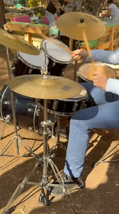 A drummer playing a kit outdoors among the pine trees