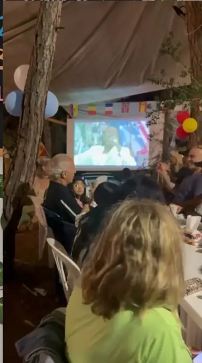 People at tables under string lights watching the World Cup final on an outdoor screen